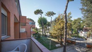A balcony with a white chair, a small garden with trees, and a view of the street. - Appartamento Aurelia (Comacchio)