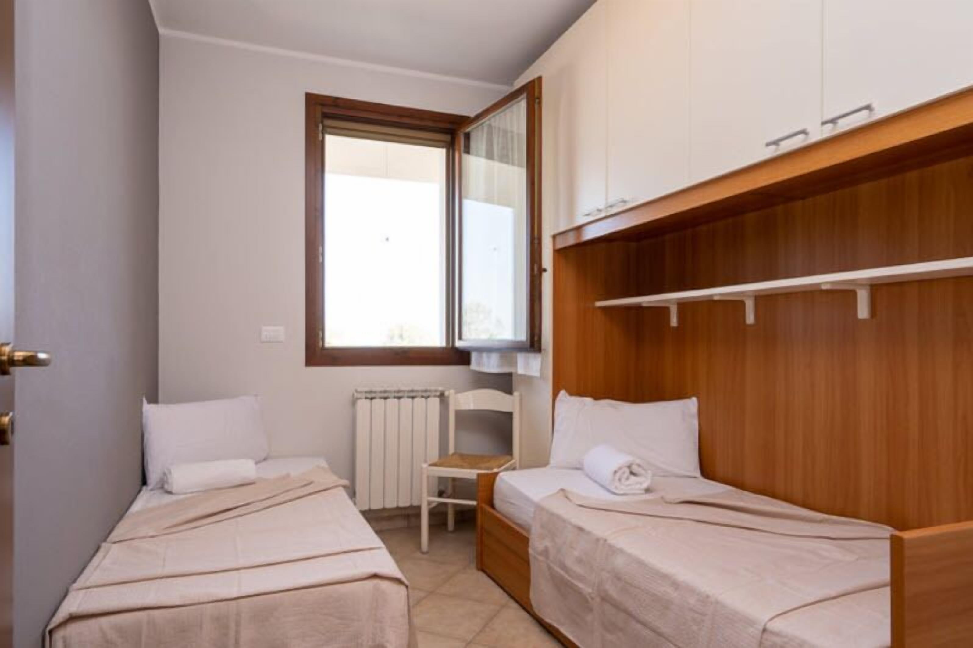 A small, clean room with two single beds, a wooden wardrobe, a radiator, and a window.