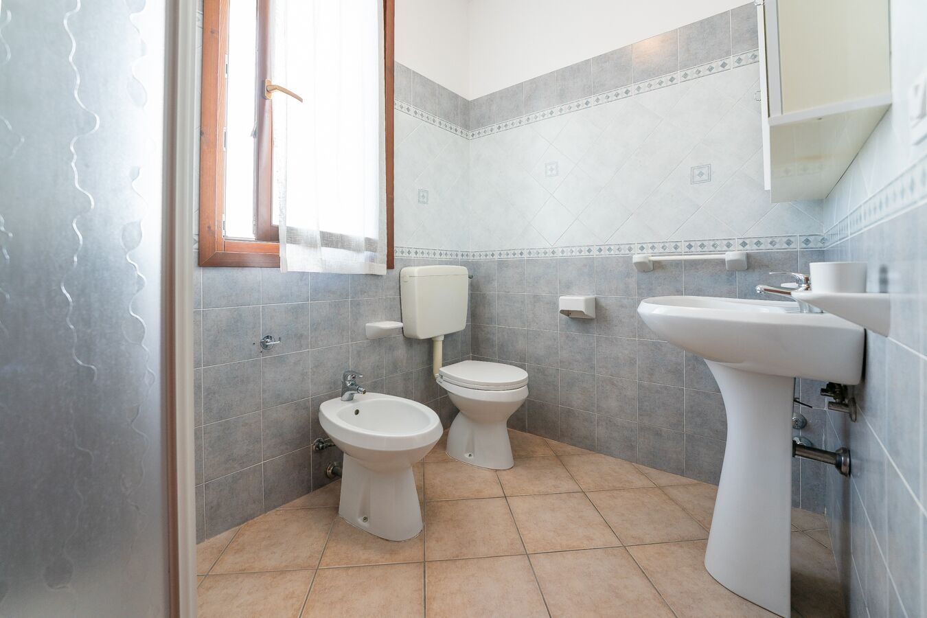 A bathroom with two toilets, a bidet, a sink, and a window with curtains.
