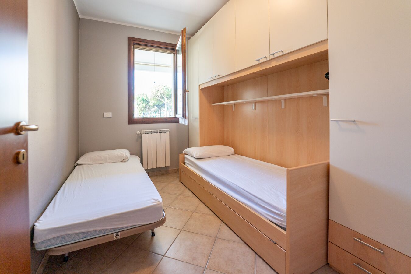 A small, well-lit room with two single beds, a wooden wardrobe, and a radiator.