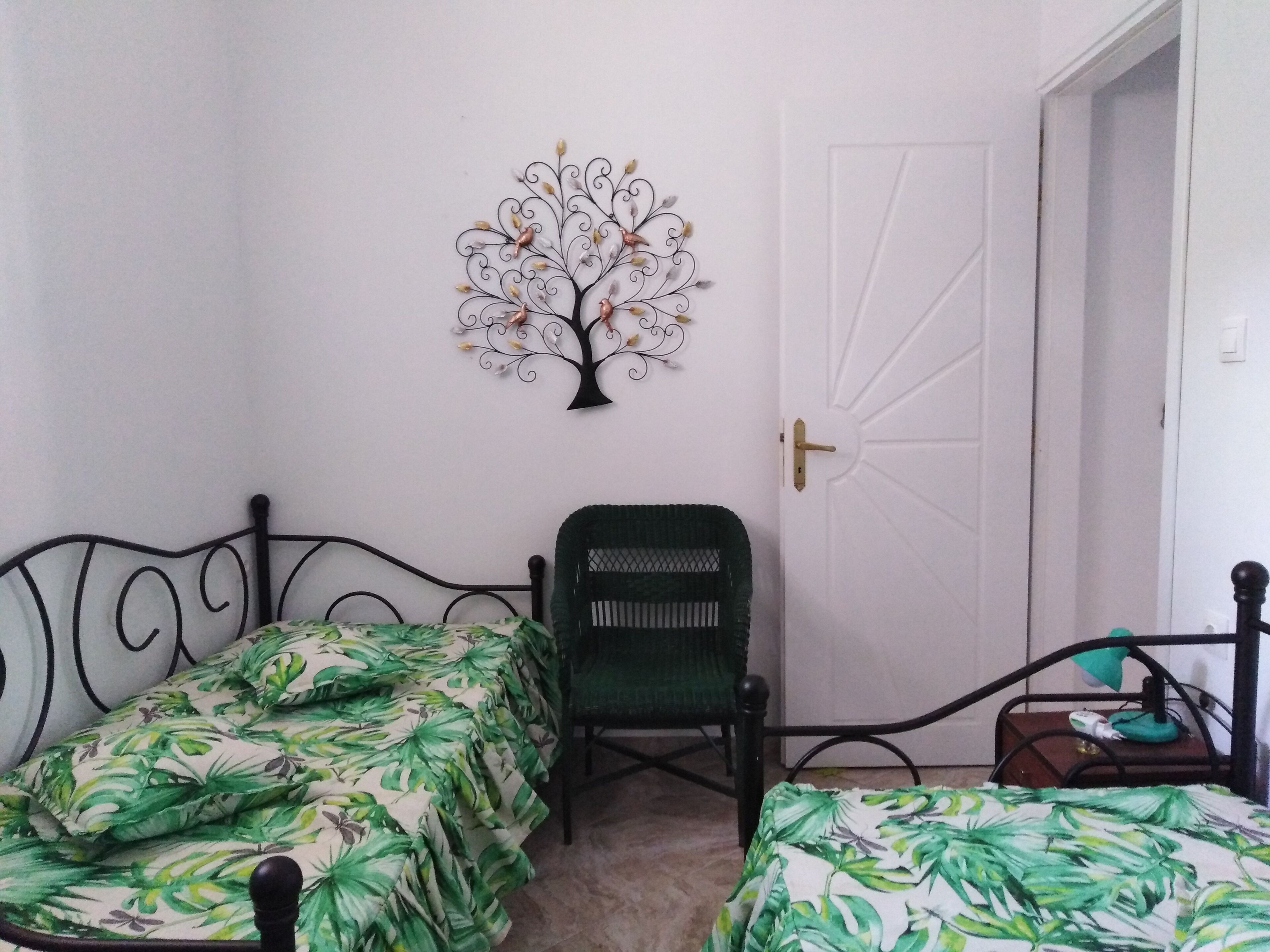 A bedroom with two single beds, a green chair, a decorative metal tree, and a white door.