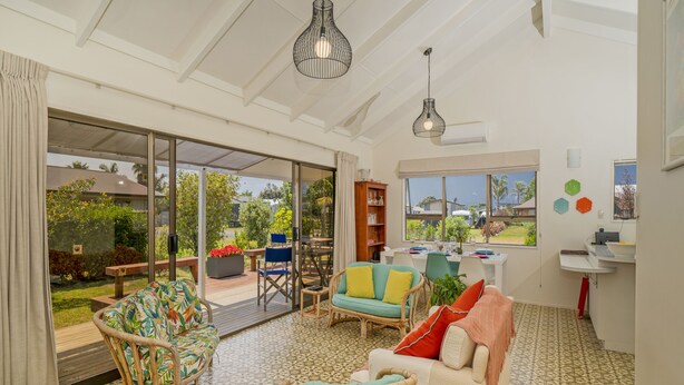 A spacious living area with a large sliding glass door leading to a patio, wicker furniture, and a kitchen area visible through the glass. - Bay Coast Happy Place - Cooks Beach Holiday Home (Cooks Beach)