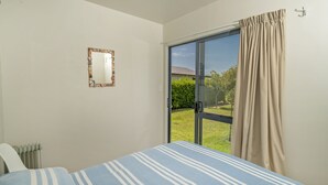 A bedroom with a bed, a window with curtains, a radiator, and a mirror on the wall. - Bay Coast Happy Place - Cooks Beach Holiday Home (Cooks Beach)