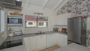 A modern kitchen with white cabinets, a stainless steel refrigerator, a microwave, and a window with a view of a house. - Bay Coast Happy Place - Cooks Beach Holiday Home (Cooks Beach)