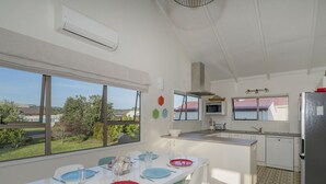 A modern kitchen with white cabinetry, a central island, and a dining area with a table set for two. - Bay Coast Happy Place - Cooks Beach Holiday Home (Cooks Beach)