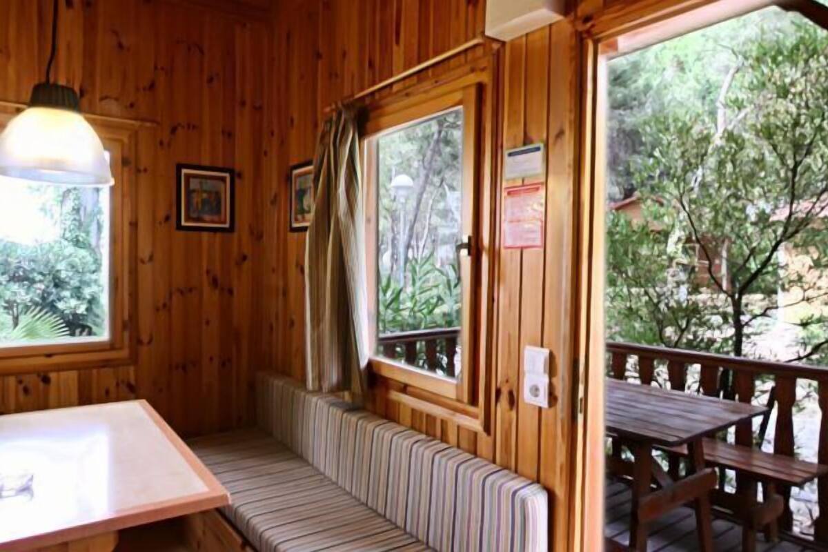A wooden cabin with a dining table, bench seating, and large windows offering a view of the outdoors.