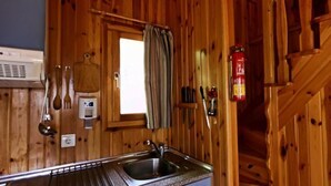 Innenansicht einer Holzhütte mit Küchenbereich, inklusive Spüle, Fenster mit Vorhang und an der Wand montiertem Feuerlöscher.
