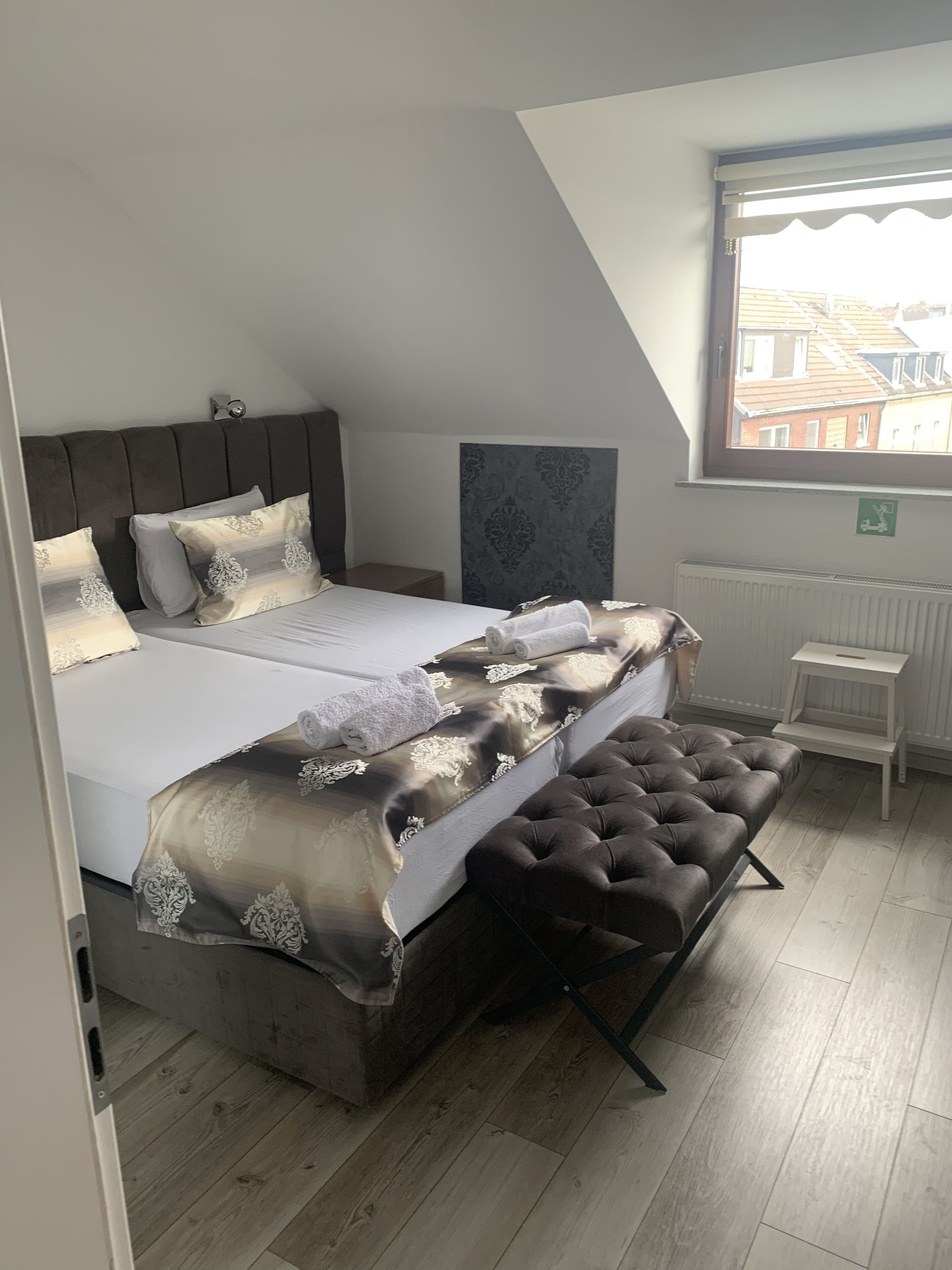 A bedroom with a large bed, a headboard, a window with a view of buildings, a radiator, and a small table.