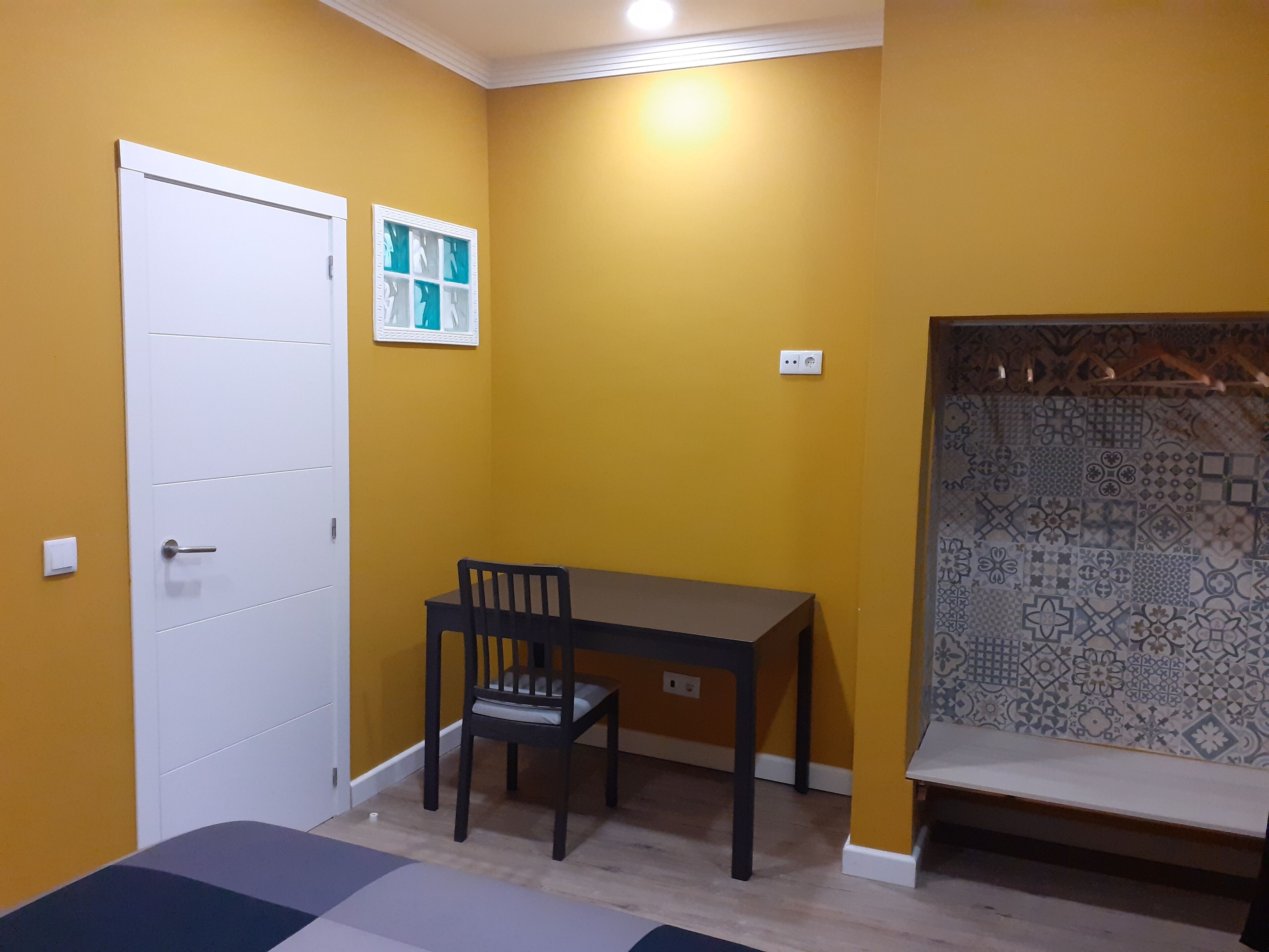 Habitació amb parets grogues, una porta blanca, una taula petita amb una cadira i un mur amb un disseny.