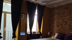 Un dormitorio con cama, ventana con cortinas, espejo y pared de piedra.