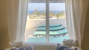 Habitació amb una finestra gran, cortines blanques, un sofà blanc i dos coixins estampats, i vista a una platja amb gandules.