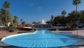 Ein Schwimmbecken mit geschwungener Kante, umgeben von Palmen und einem Gebäude mit Kegeldach im Hintergrund.
