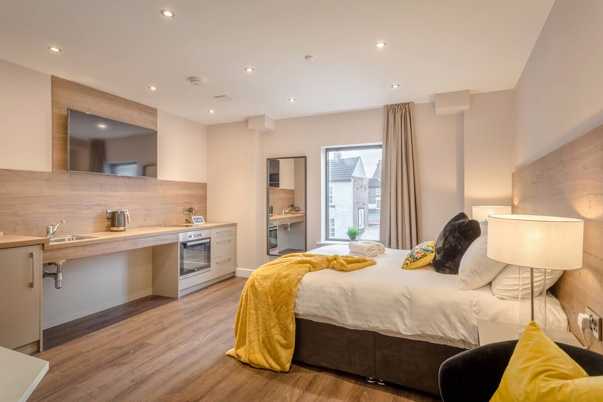Une chambre d’hôtel moderne avec un coin cuisine, un grand lit recouvert d’un plaid jaune et une vue sur la ville par la fenêtre.