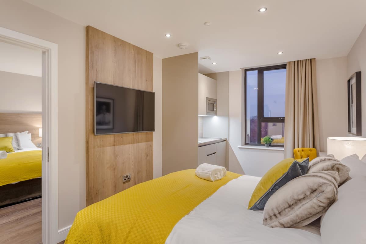 Une chambre d’hôtel moderne dotée d’un grand lit, d’un téléviseur à écran plat fixé au mur, d’une petite kitchenette et d’une fenêtre avec des rideaux.