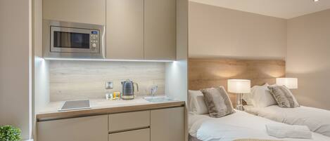 A modern hotel room with a kitchenette, a bed with two pillows, bedside lamps, and a bedside table.