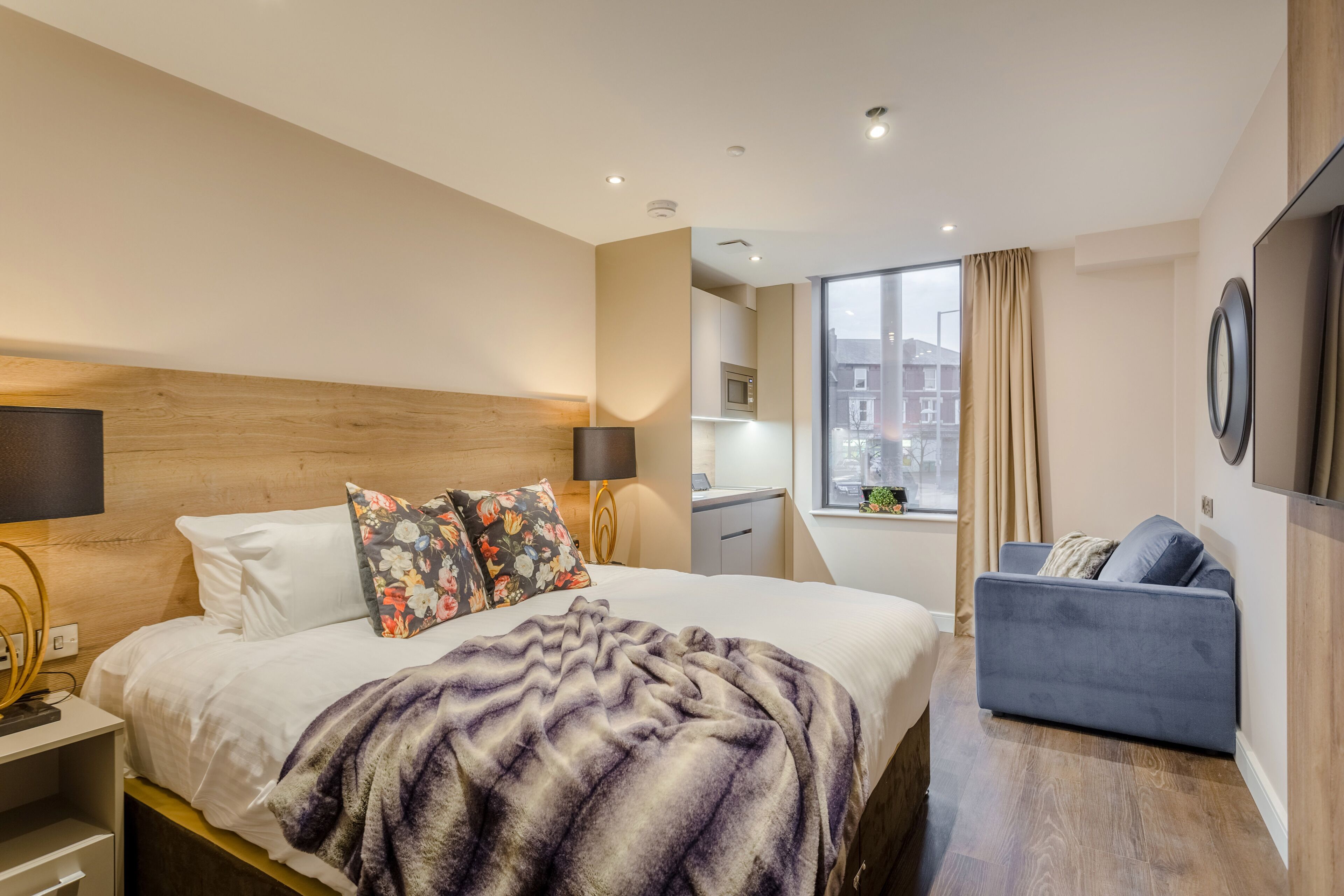 Habitación de hotel con cama, sofá azul, cabecera de madera, ventana con cortinas y vista a la ciudad.