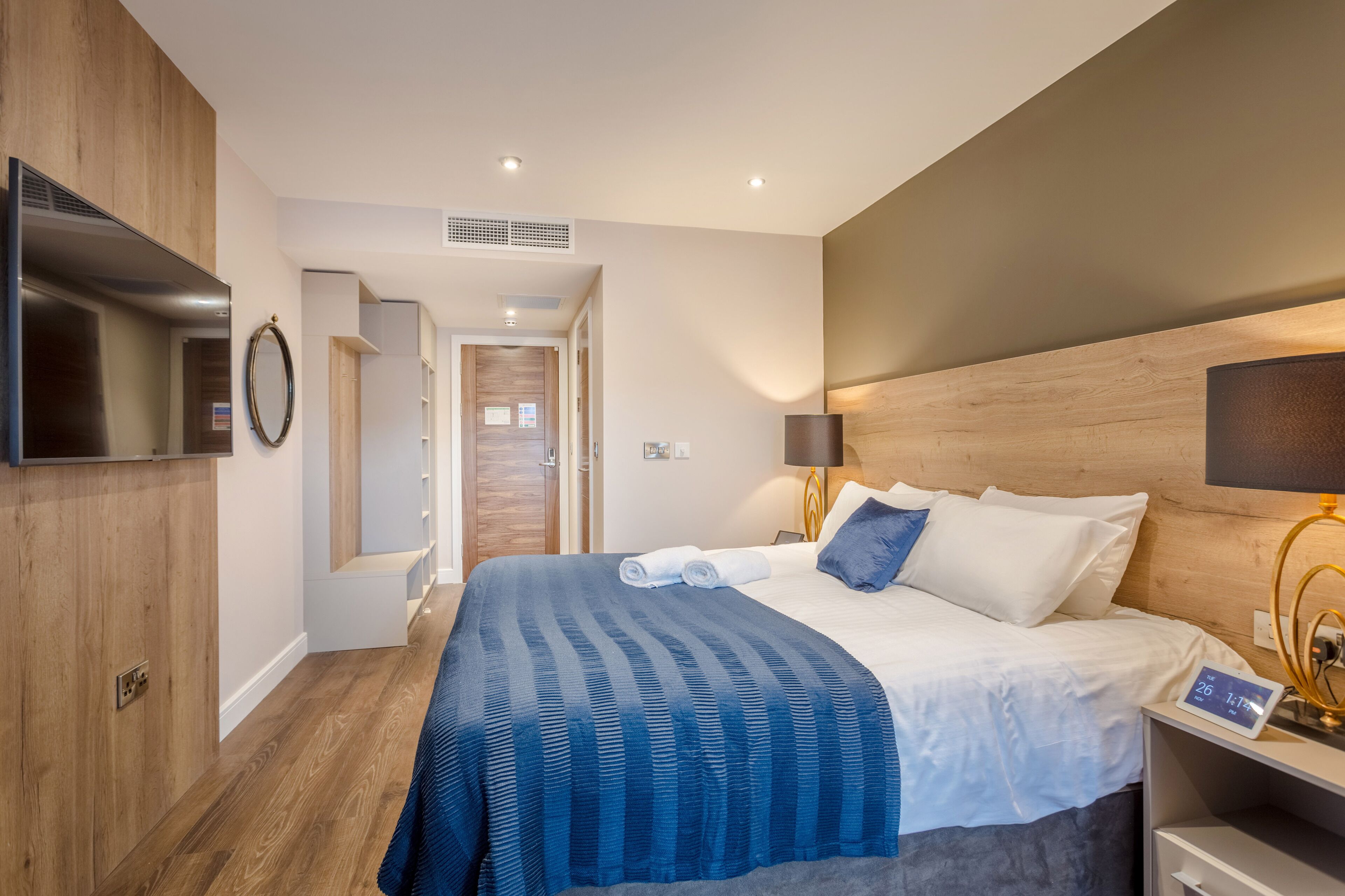 A modern hotel room with a large bed, a flat-screen TV, a wooden headboard, and a bedside table with a lamp.