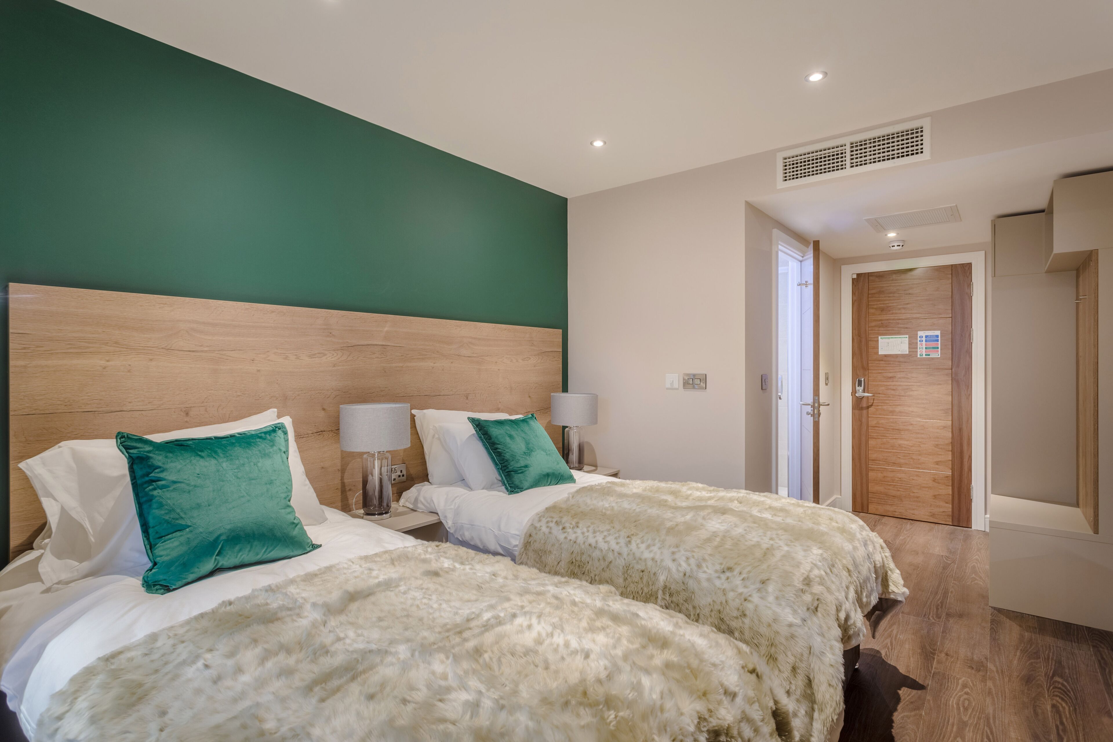 Una camera d'albergo con due letti, pareti verdi, una testiera in legno, biancheria da letto bianca e una porta che conduce a un'altra stanza.