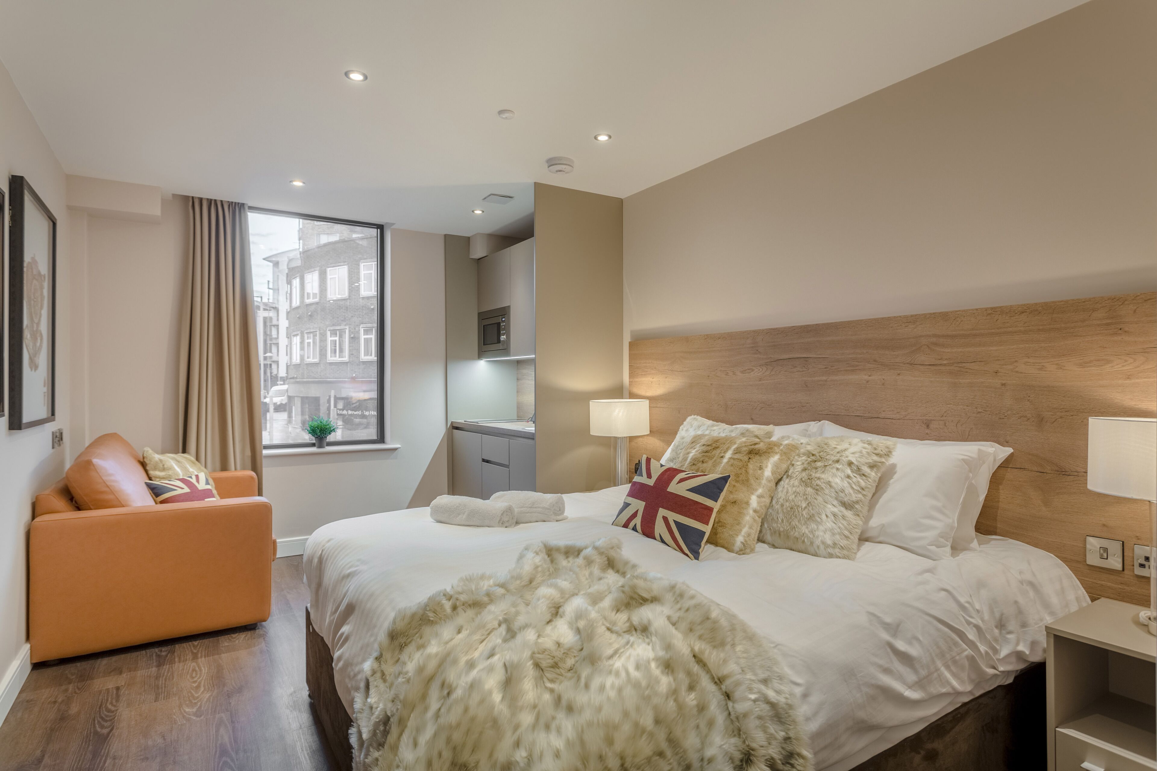 Habitación de hotel moderna con una cama grande, un sofá, un cabecero de madera, una ventana con cortinas y vistas a edificios.