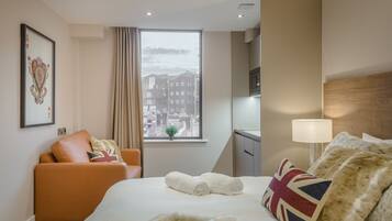 A hotel room with a bed, a sofa, a window with a city view, a lamp, and a framed picture on the wall.