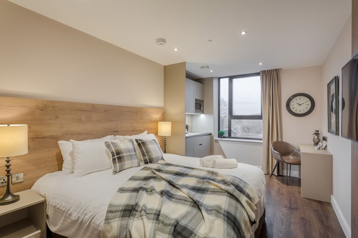 Une chambre d’hôtel avec une tête de lit en bois, un lit avec du linge de maison blanc, une couverture à carreaux, un bureau avec une chaise, une horloge et une fenêtre avec des rideaux.