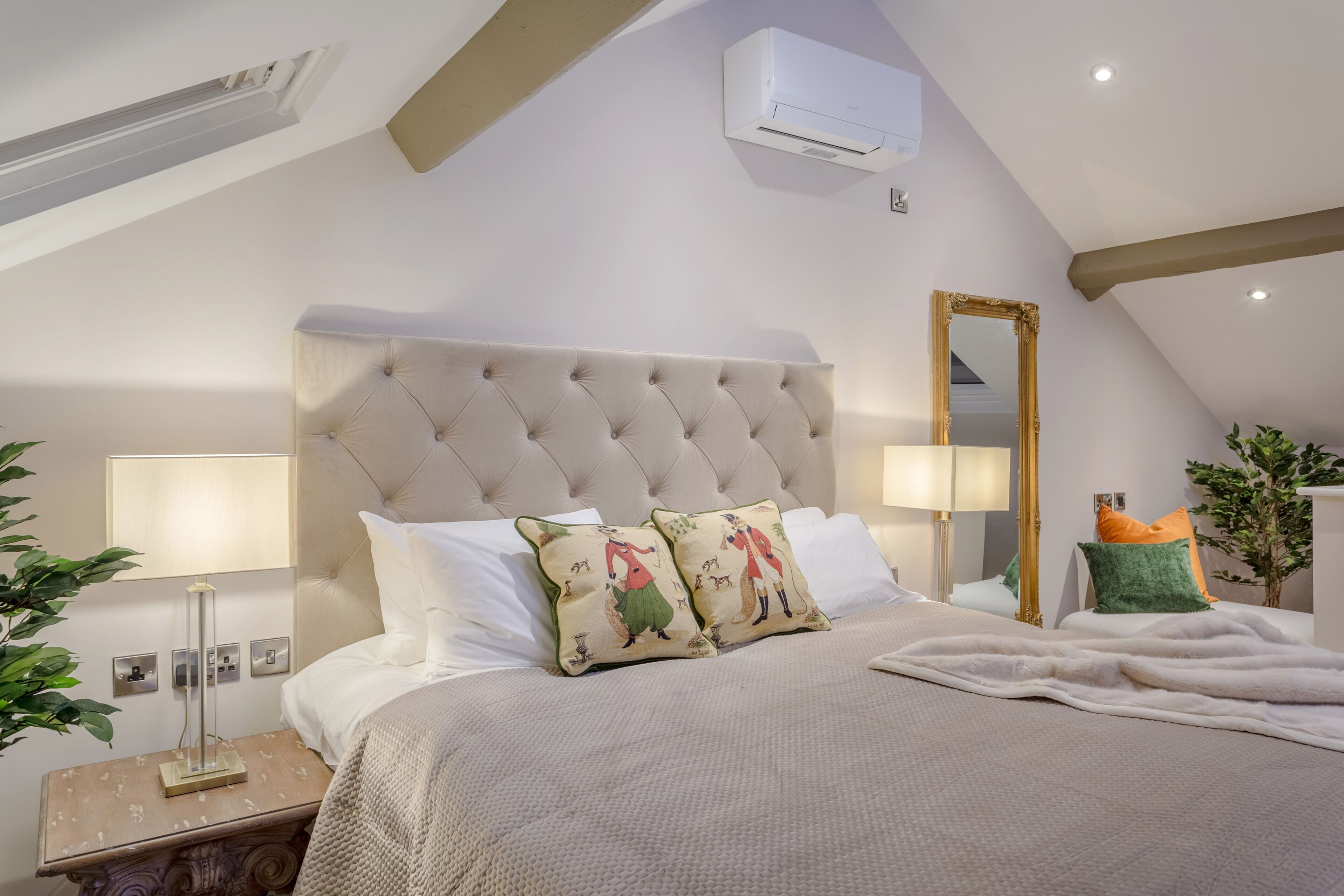 A bedroom with a tufted headboard, white bedding, decorative pillows, a bedside lamp, and an air conditioning unit.