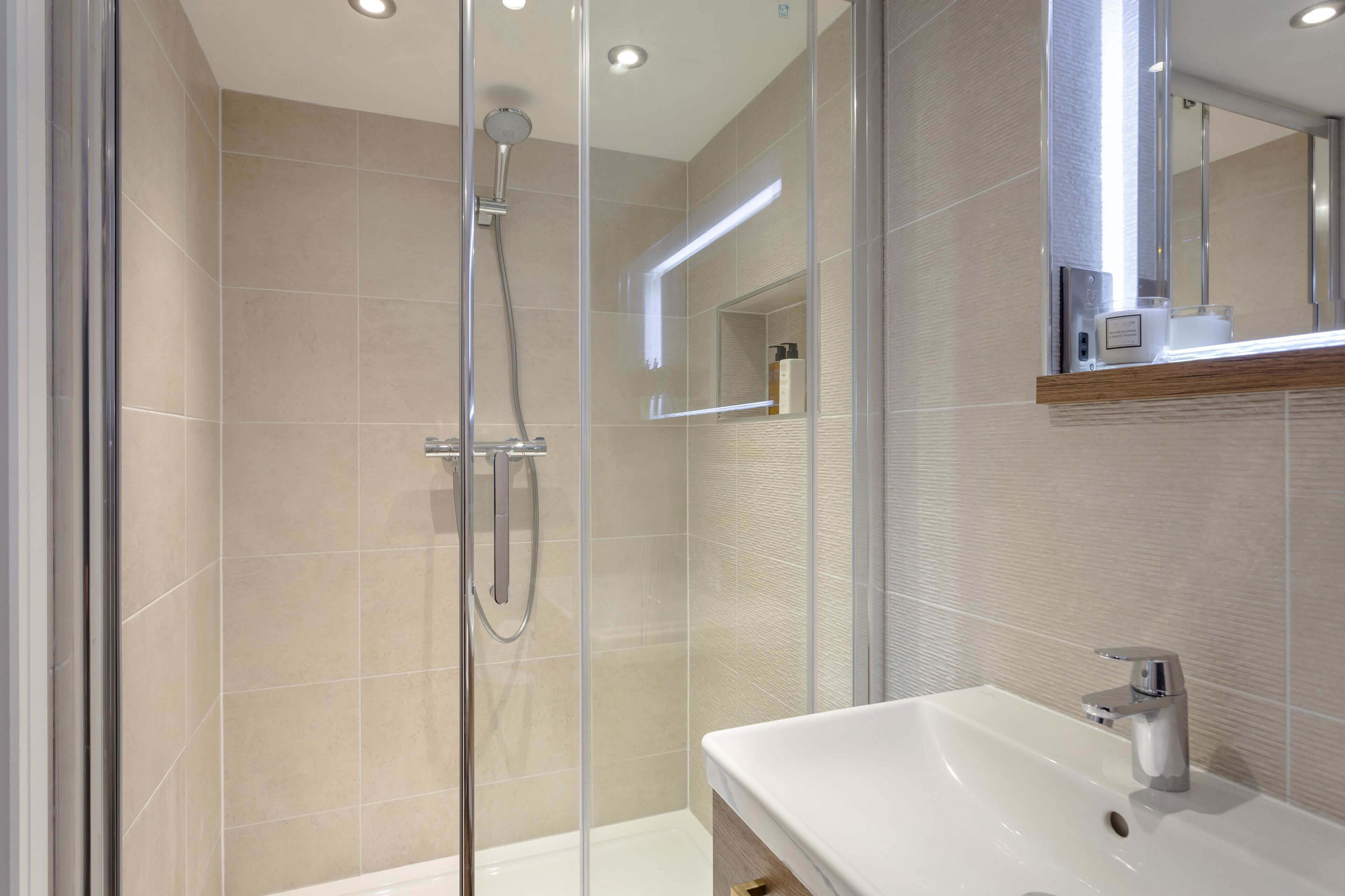 Un baño moderno con ducha de vidrio, lavamanos blanco y espejo con repisa de madera.