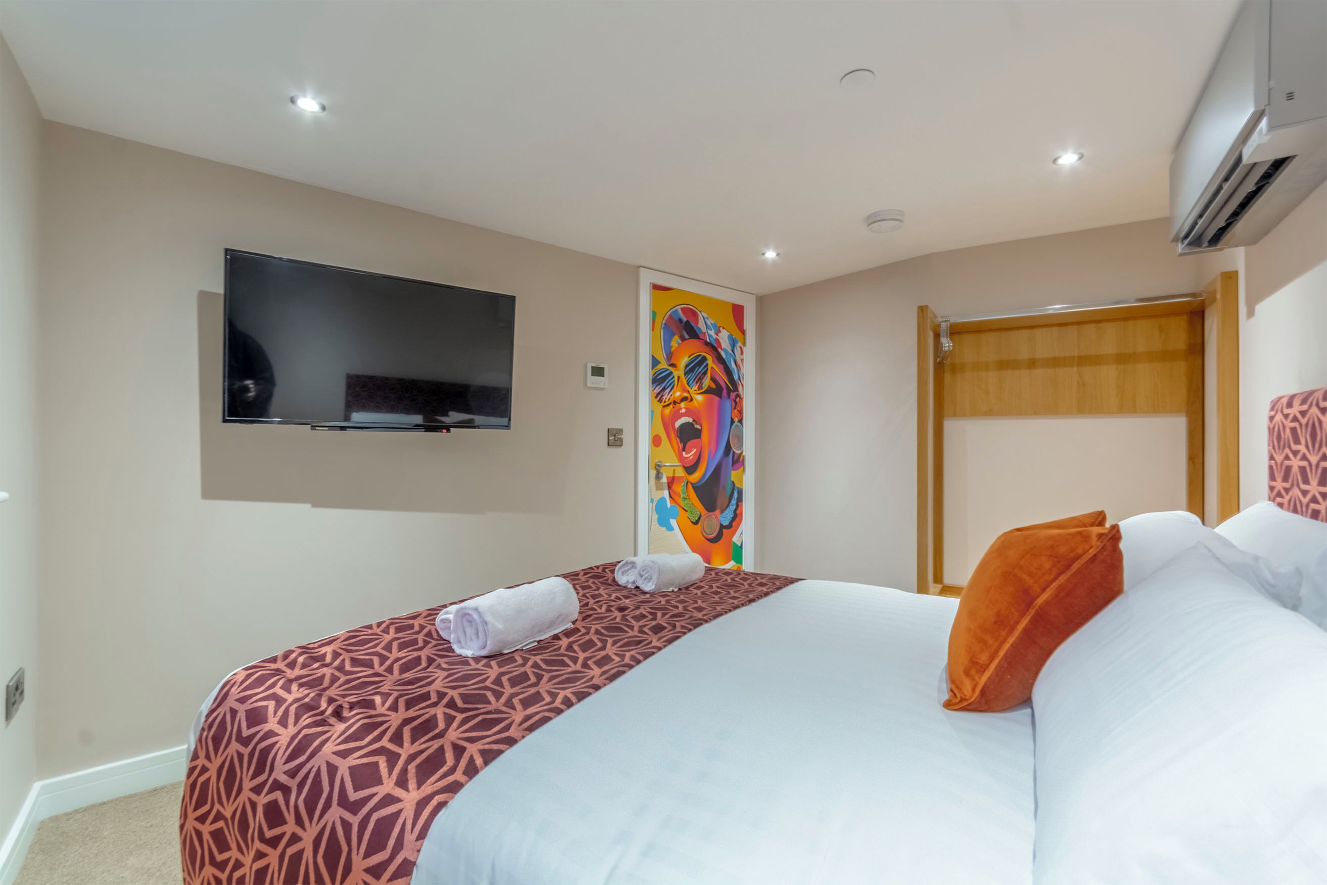 A hotel room with a large bed, a flat-screen TV mounted on the wall, a colorful artwork on the door, and a wooden headboard.