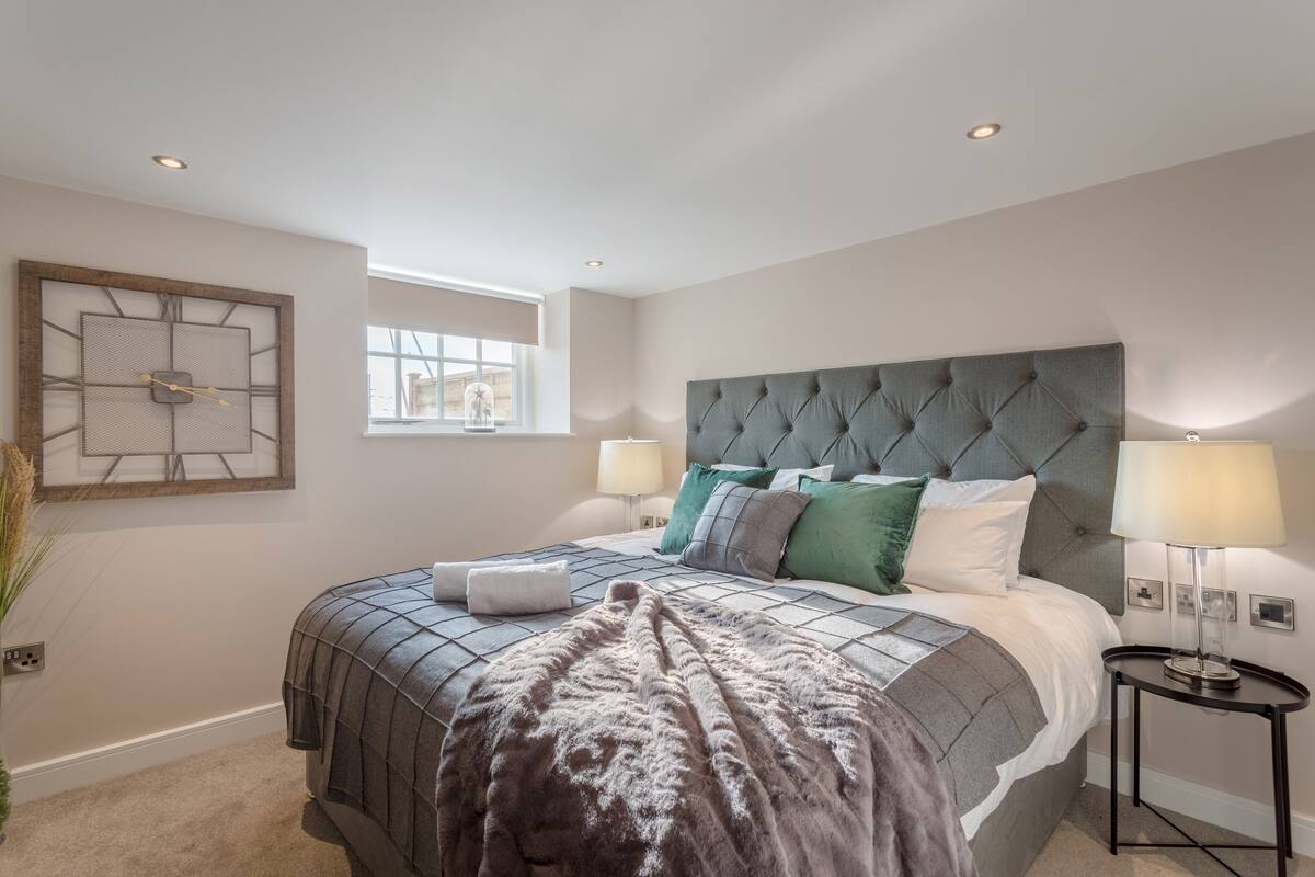 A bedroom with a tufted headboard, a large bed with a grey and white checkered bedspread, multiple pillows, a bedside table with a lamp, and a window with a view of the outside.