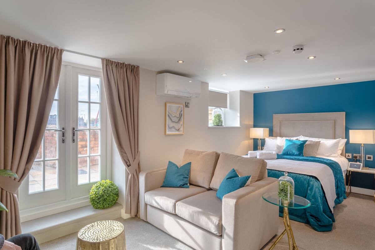 A modern hotel room with a beige sofa, a bed with blue bedding, and a glass coffee table.