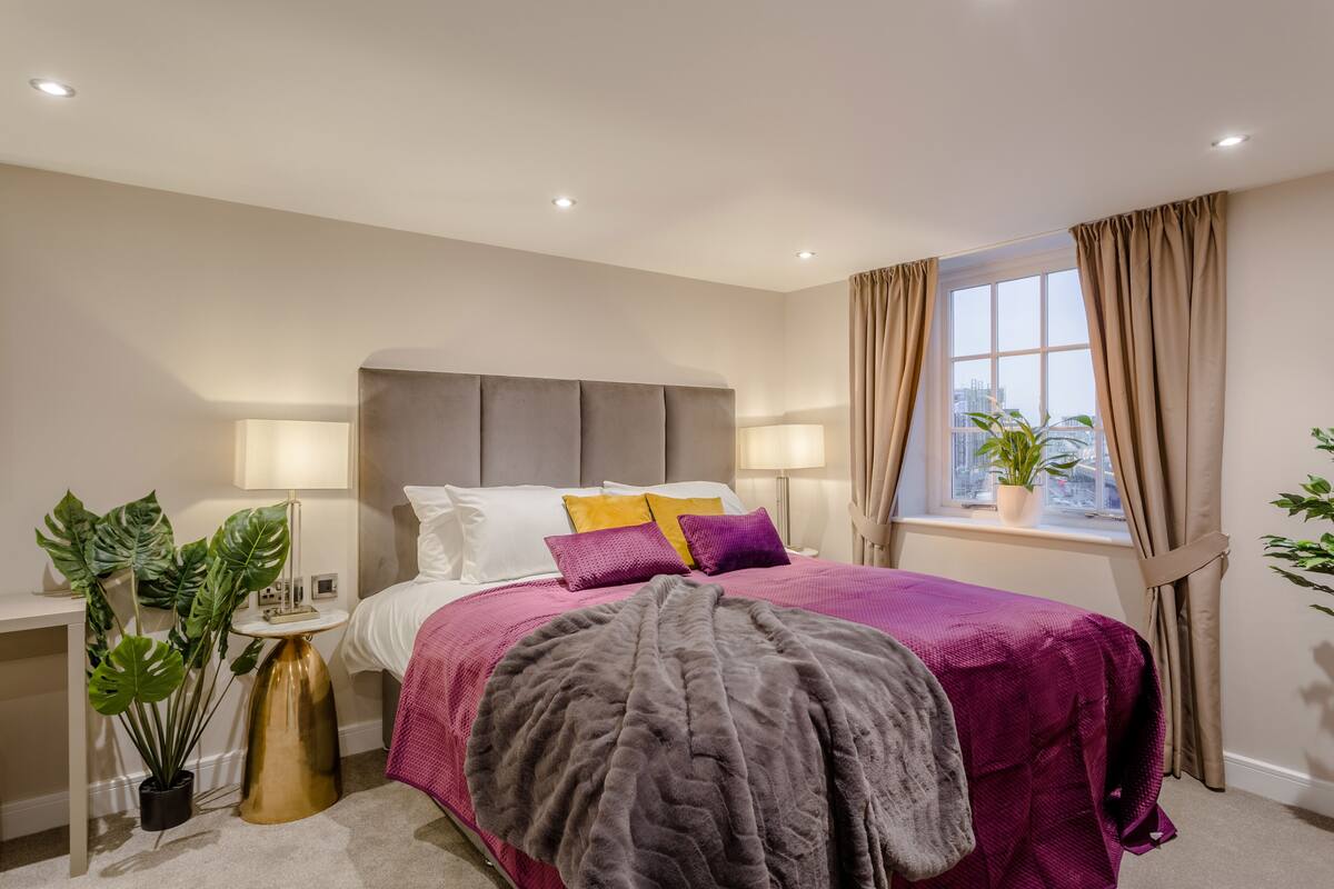 A modern bedroom with a large bed, bedside lamps, a window with curtains, and a potted plant.
