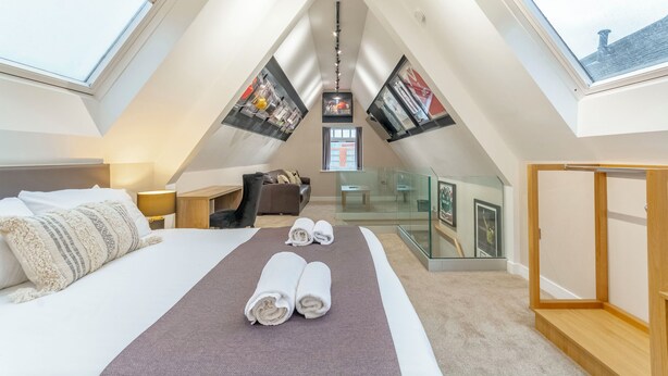 Una habitación con techos altos, tragaluces, una cama con toallas enrolladas, un separador de vidrio y un armario de madera.
