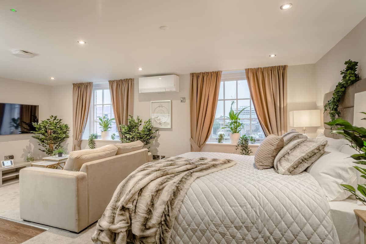 A modern hotel room with a large bed, a sofa, a TV, and potted plants.