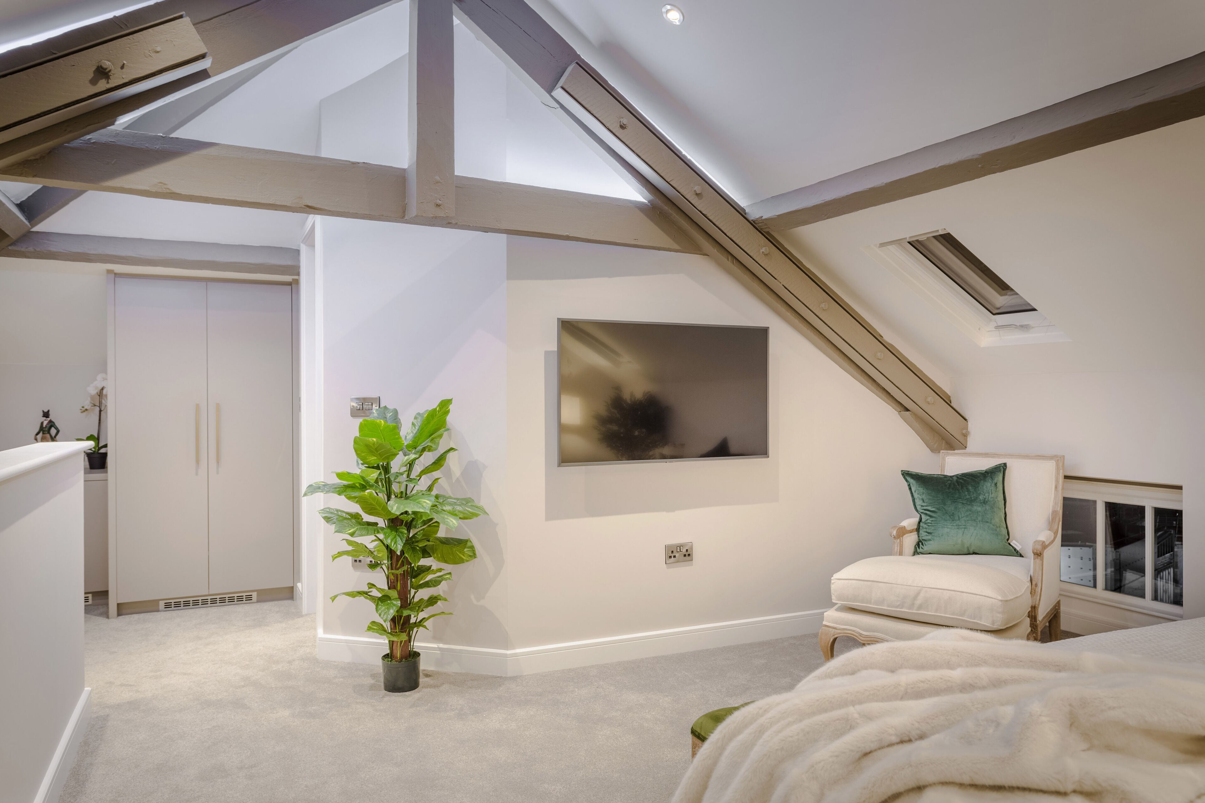 A modern bedroom with a sloped ceiling, a large window, a bed with a white blanket, a chair with a green pillow, and a potted plant.