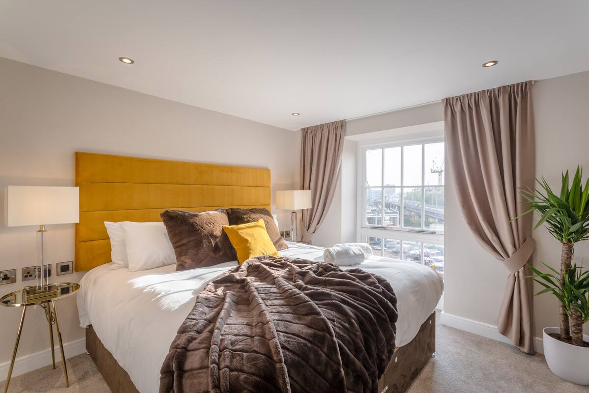 A hotel room with a large bed, a headboard, two bedside lamps, a window with curtains, and a potted plant.
