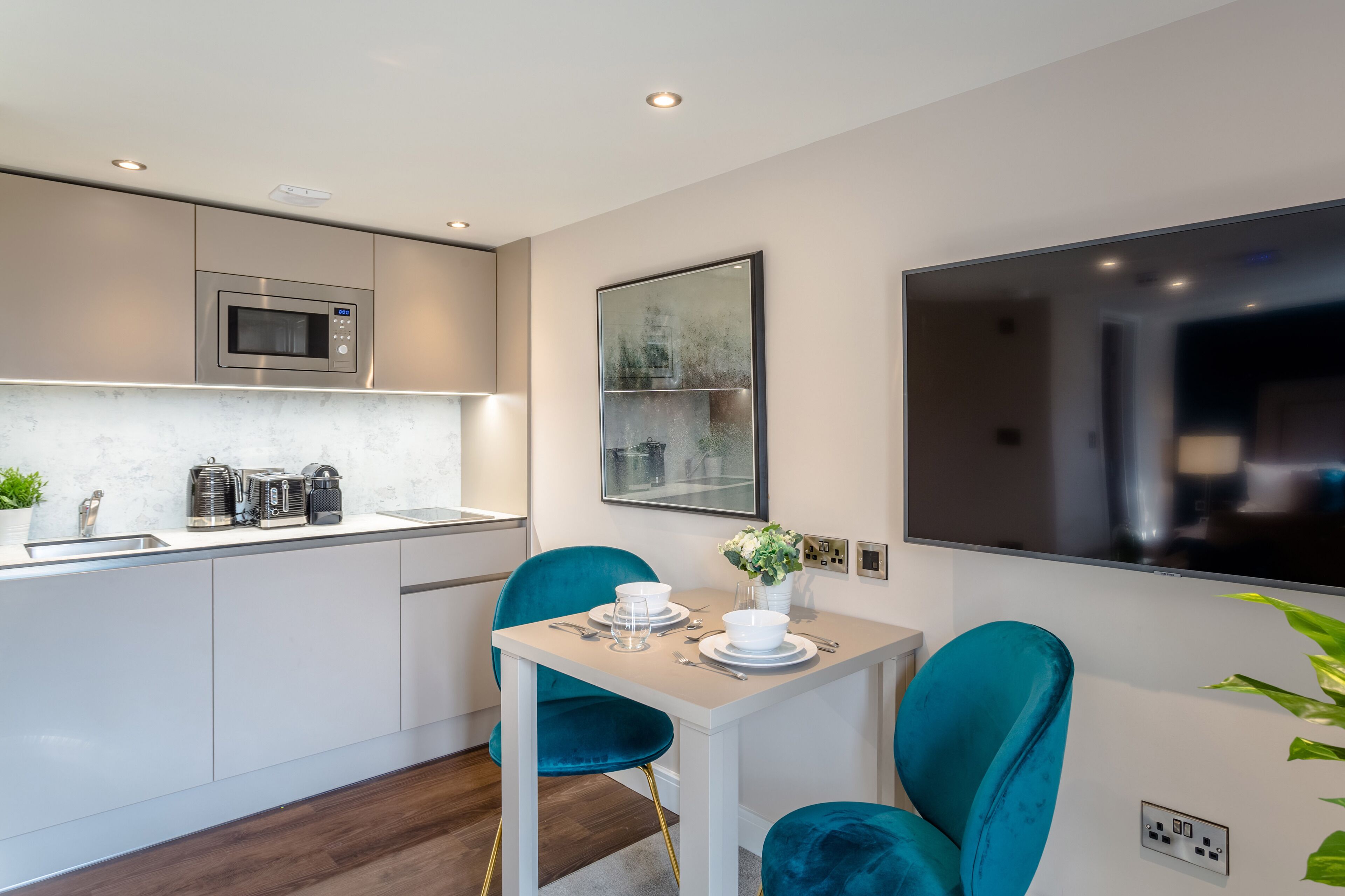 A modern kitchen with a microwave, sink, and countertop appliances. There is a dining area with a table and chairs, and a large mirror on the wall.