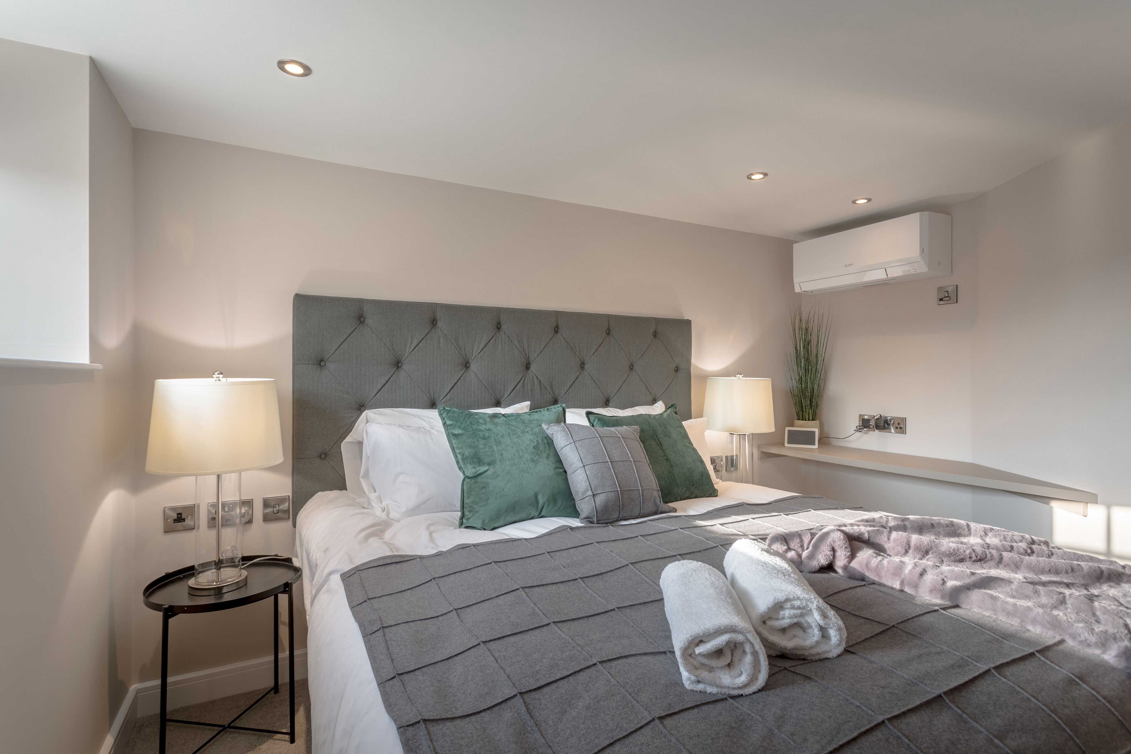 A modern hotel room with a large bed, two bedside tables, a grey headboard, and an air conditioning unit on the wall.