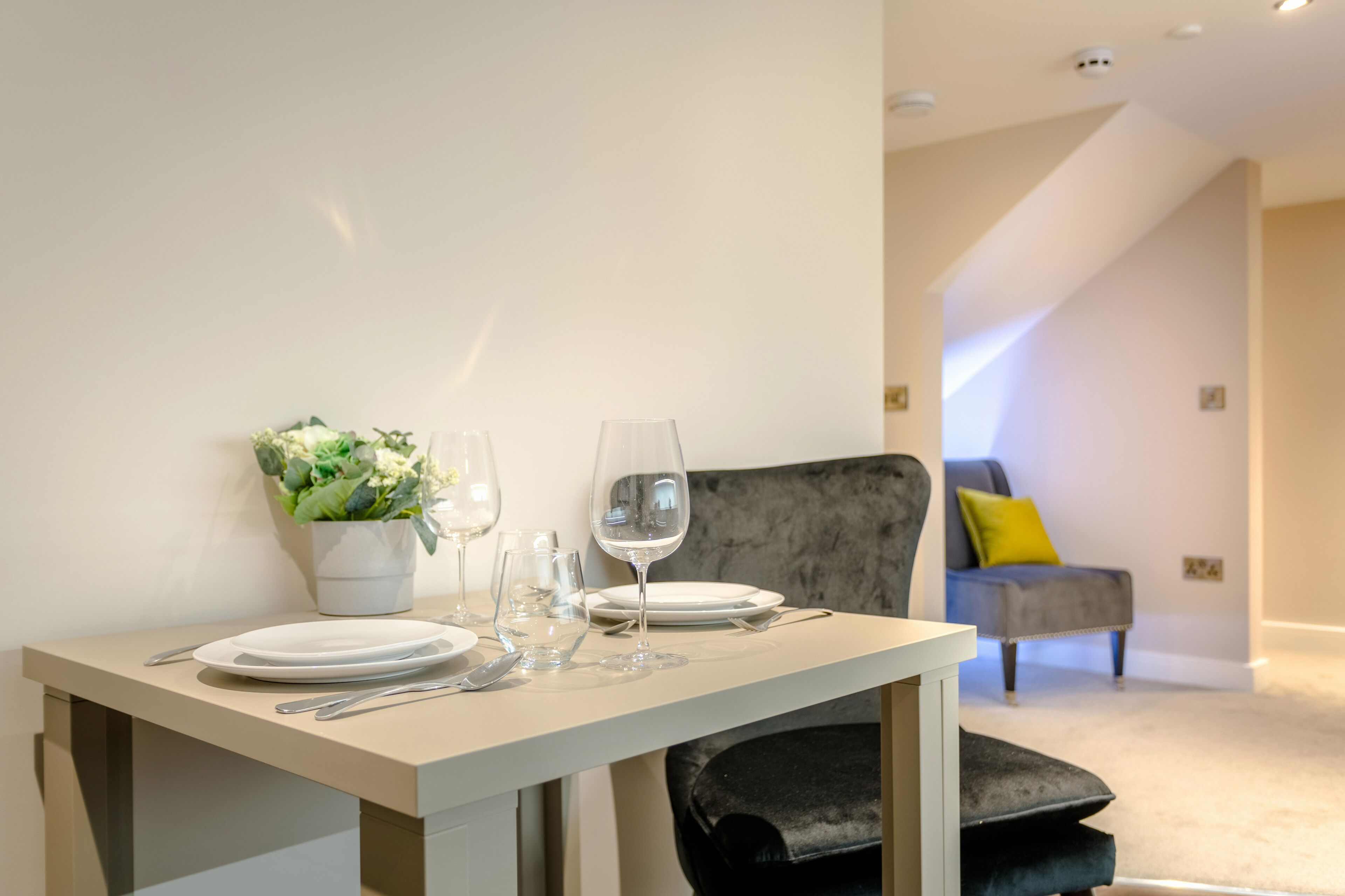 A dining area with a table set for two, a potted plant, and a grey armchair in the background.
