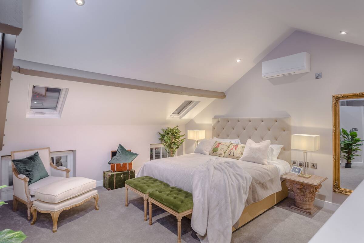 A bedroom with a large bed, a chair, a bench, and a bedside table with a lamp.