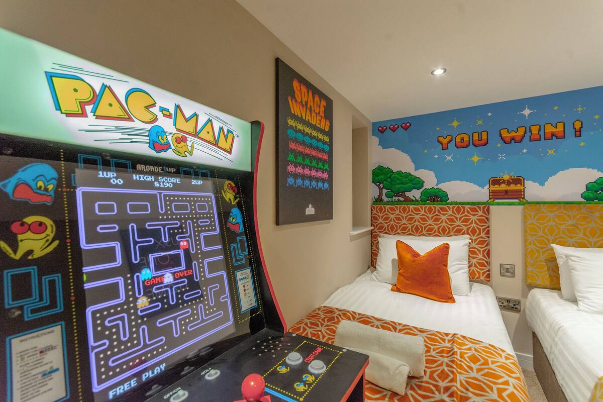 A room with a Pac-Man arcade game, a bed with orange and white bedding, and a 'YOU WIN!' wall decoration.