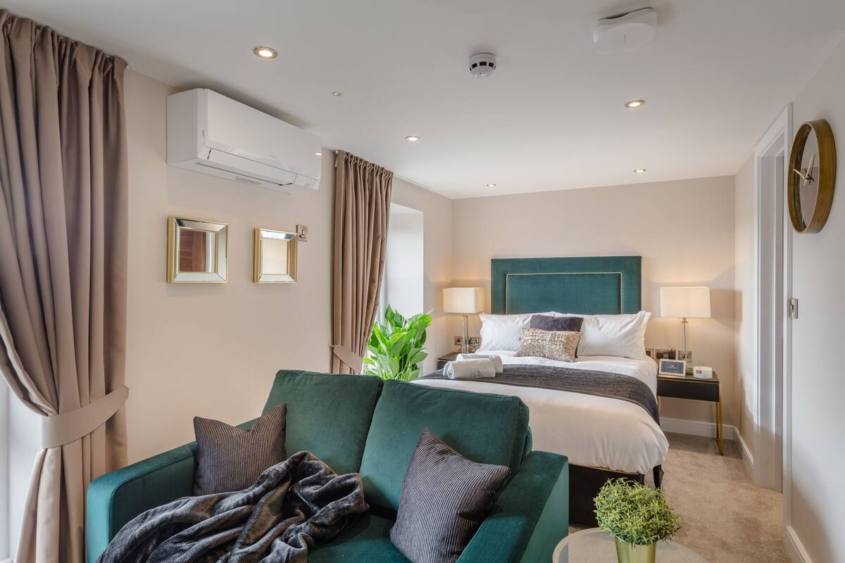 A hotel room with a bed, a green sofa, a small table with a plant, and a wall-mounted air conditioner.