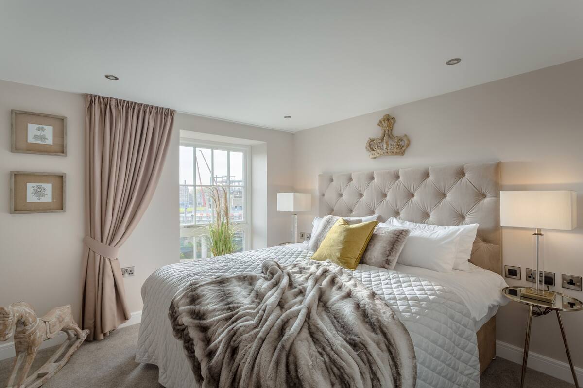 A bedroom with a tufted headboard, a large bed with a fluffy blanket, a bedside table with a lamp, and a window with curtains.