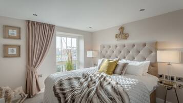 Een slaapkamer met een gestoffeerd hoofdeinde, een groot bed met een zachte deken, een nachtkastje met een lamp en een raam met gordijnen.
