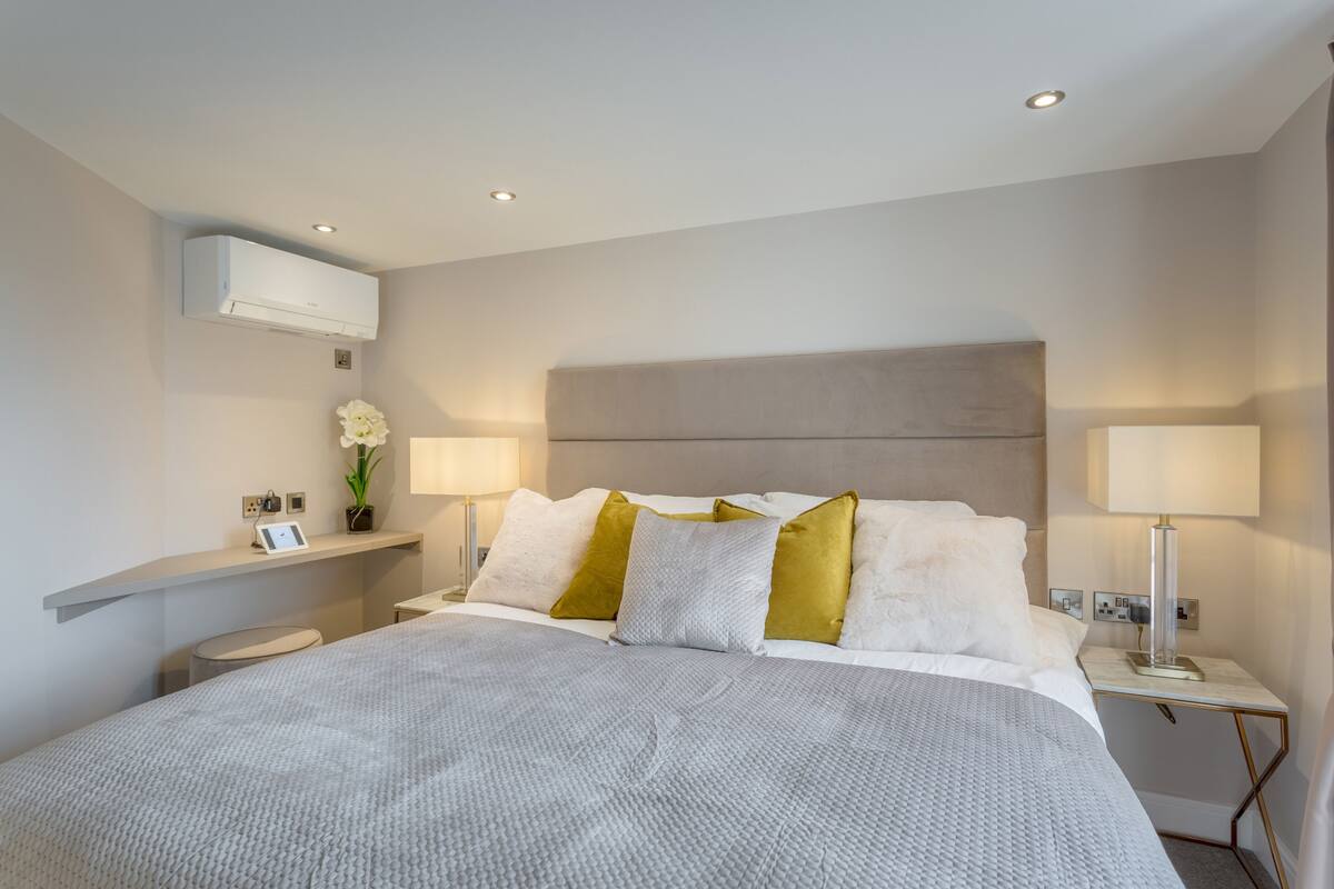 A modern hotel room with a large bed, bedside lamps, a nightstand, a small table, and an air conditioning unit.