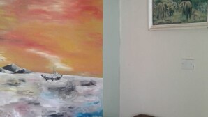 A living room with a brown sofa, a painting of a sunset over water, and a framed landscape painting on the wall. - Greenview Guest House (Kisumu)