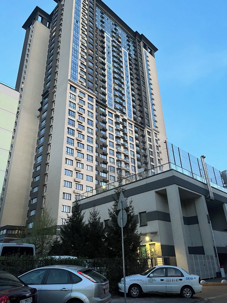 A tall residential building with multiple balconies, a parking area with cars, and a street lamp.