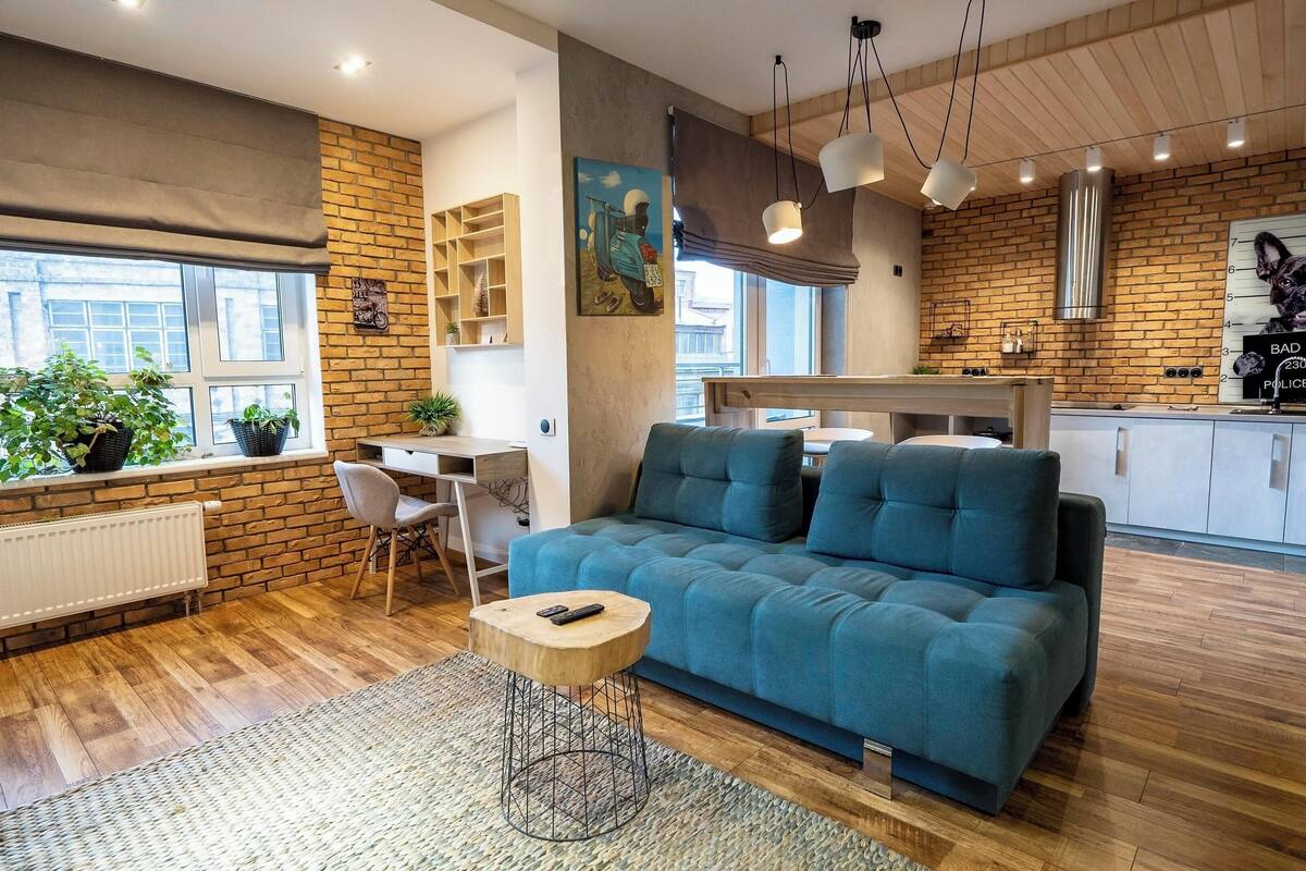 Una sala de estar moderna con un sofá azul, suelo de madera, comedor y cocina con paredes de ladrillo.
