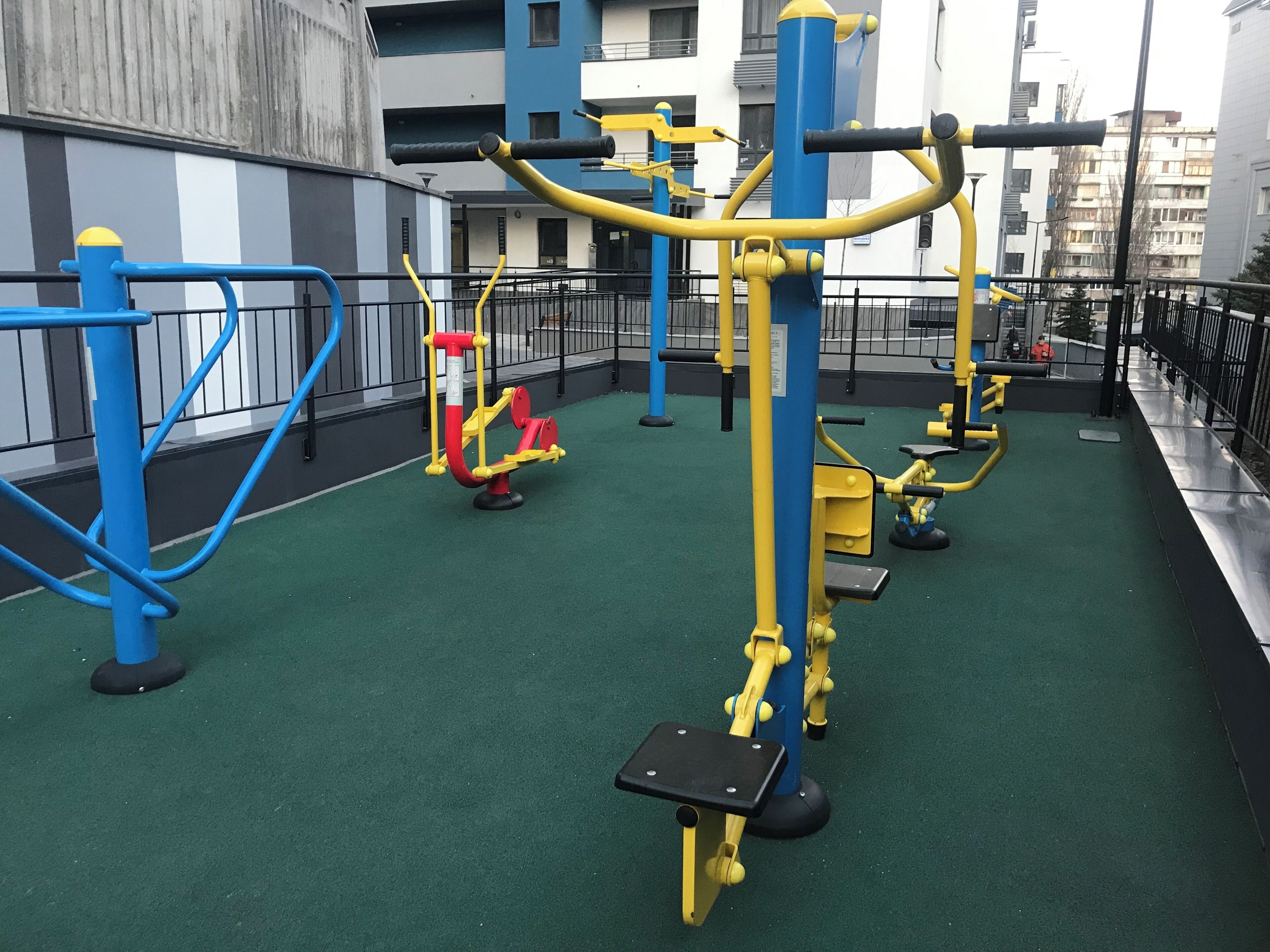 Ein Outdoor-Fitnessbereich mit Trainingsgeräten auf einer Grünfläche, umgeben von einem Metallzaun.