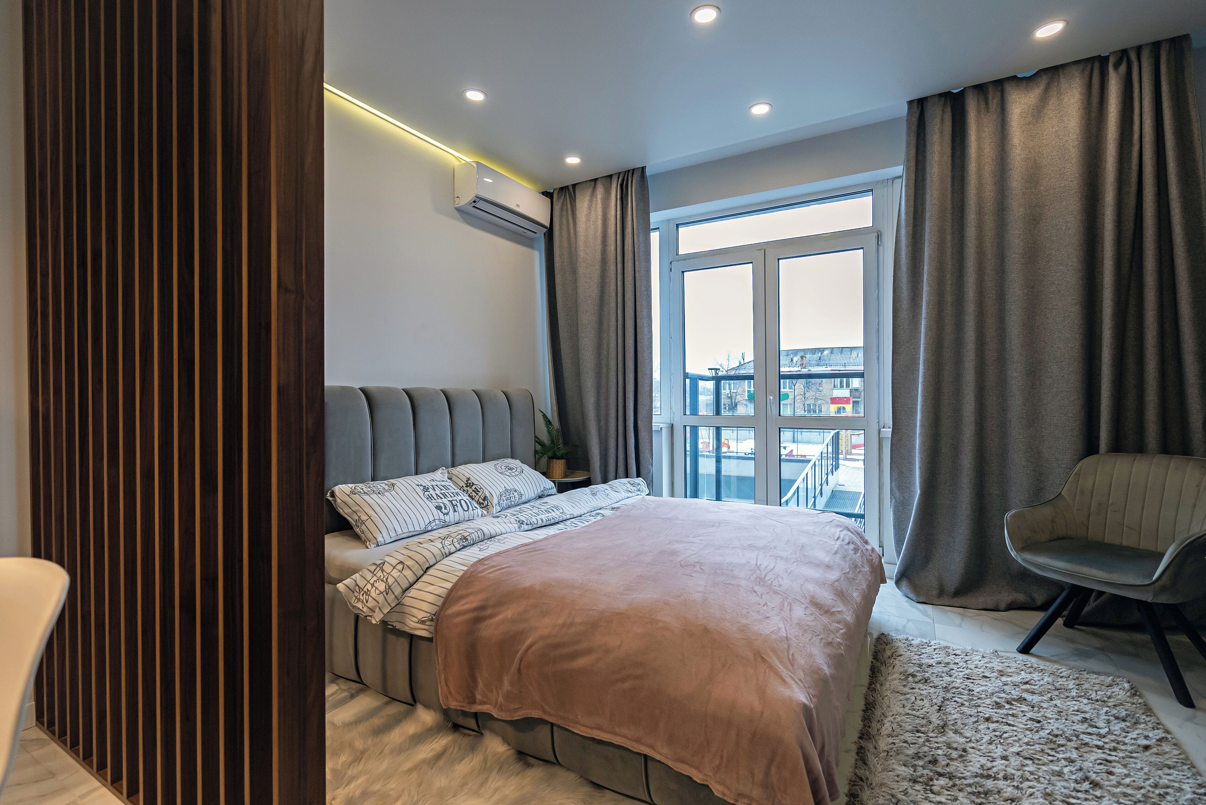 Quarto moderno com uma cama grande, uma poltrona cinza e vista para a cidade através de uma janela ampla.