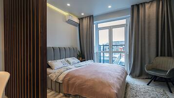 Quarto moderno com uma cama grande, uma poltrona cinza e vista para a cidade através de uma janela ampla.
